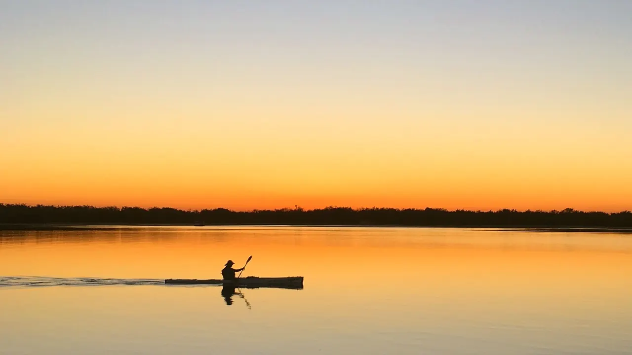 A lone kayaker silhouetted on calm golden water at dusk in Biggera Waters, Australia.