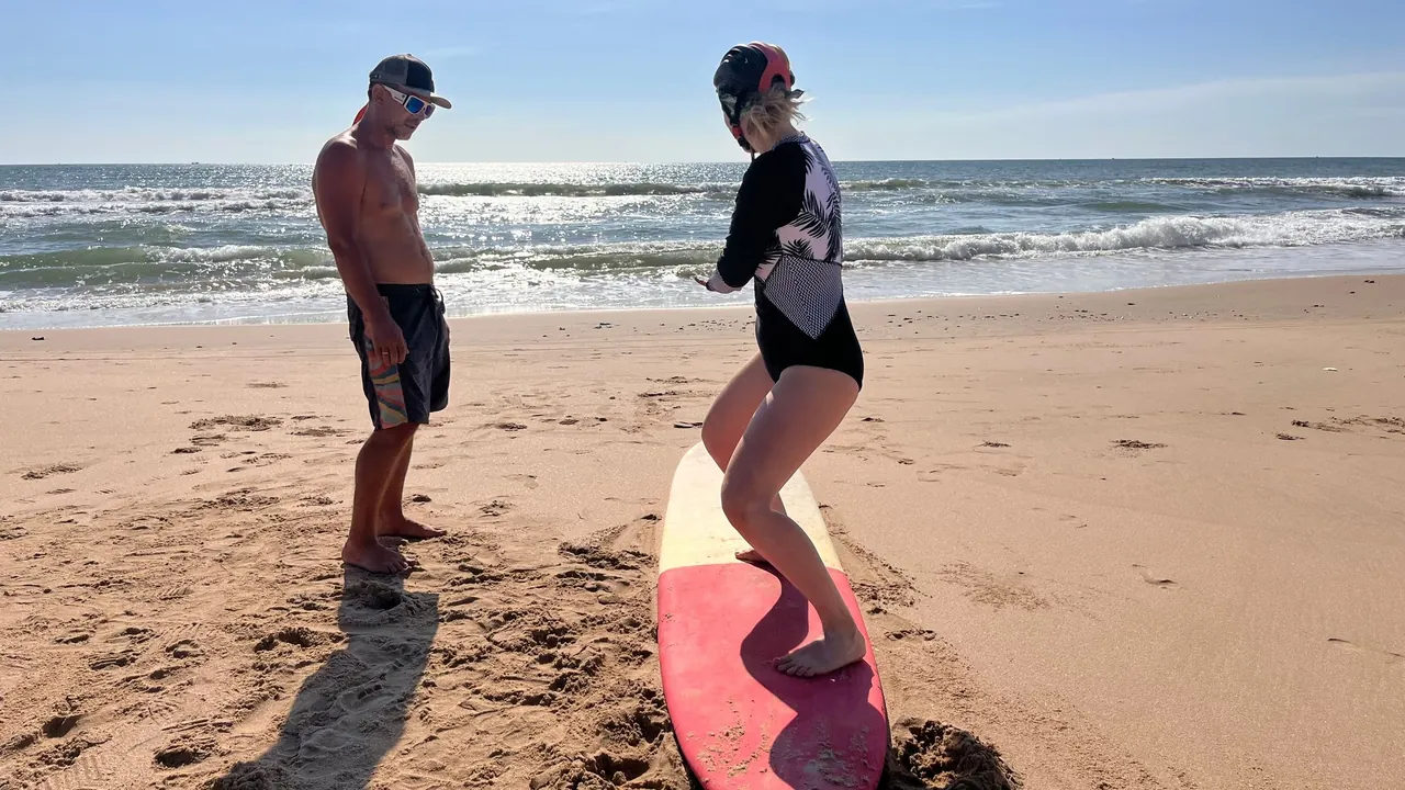 A beginner stands on a pink surfboard on the sand while an instructor guides her.