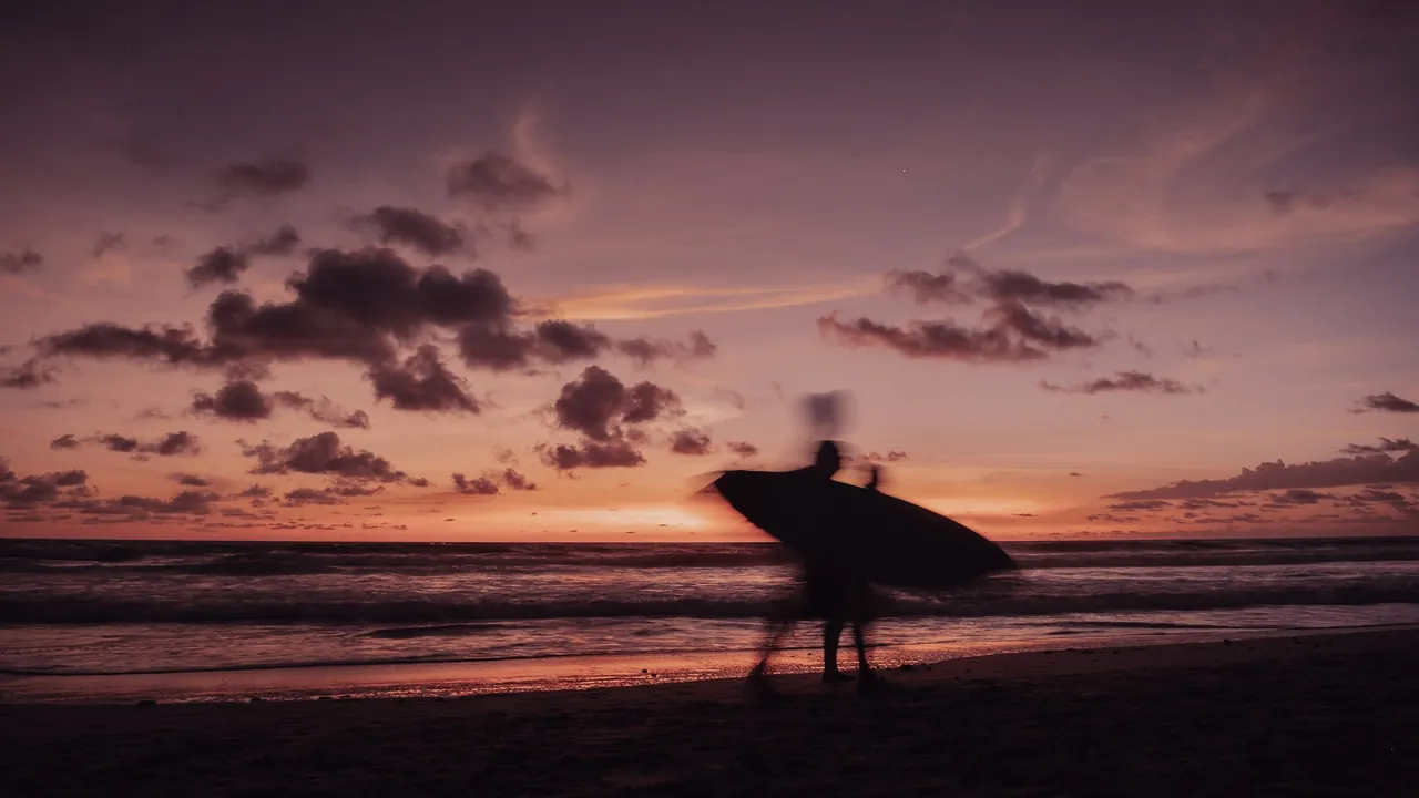 A surfer with a board silhouetted against a violet and orange sunset at Santa Teresa Beach.