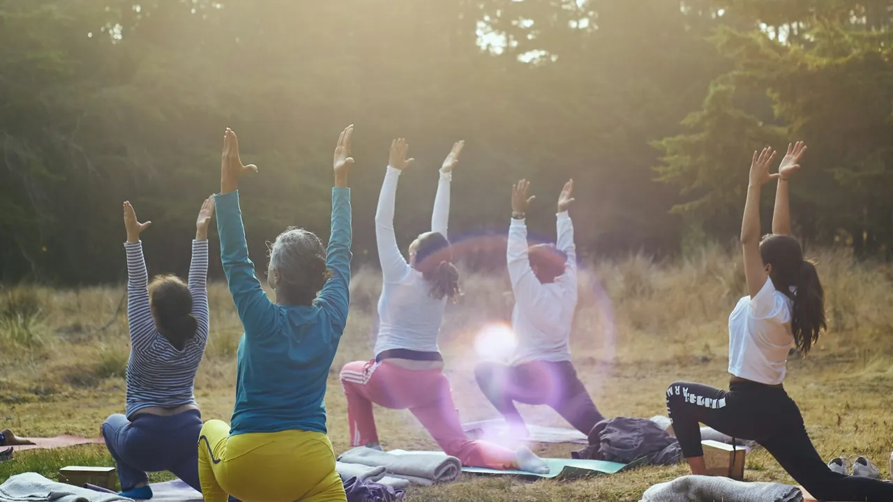 A group practicing yoga on blankets in a grassy clearing with warm sunlight filtering through trees.